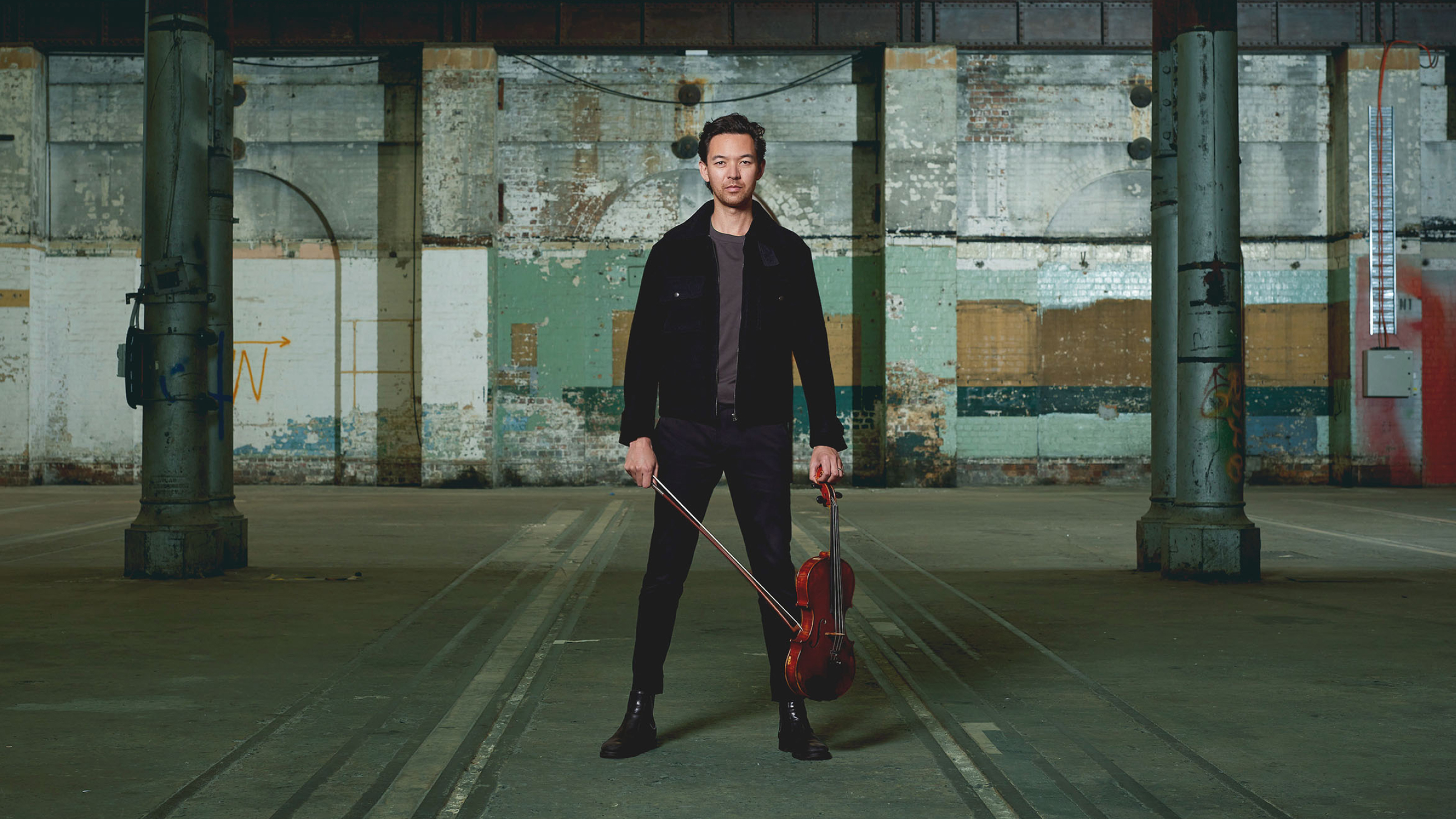A man standing in an empty warehouse holding a violin and bow in each hand down by his sides.