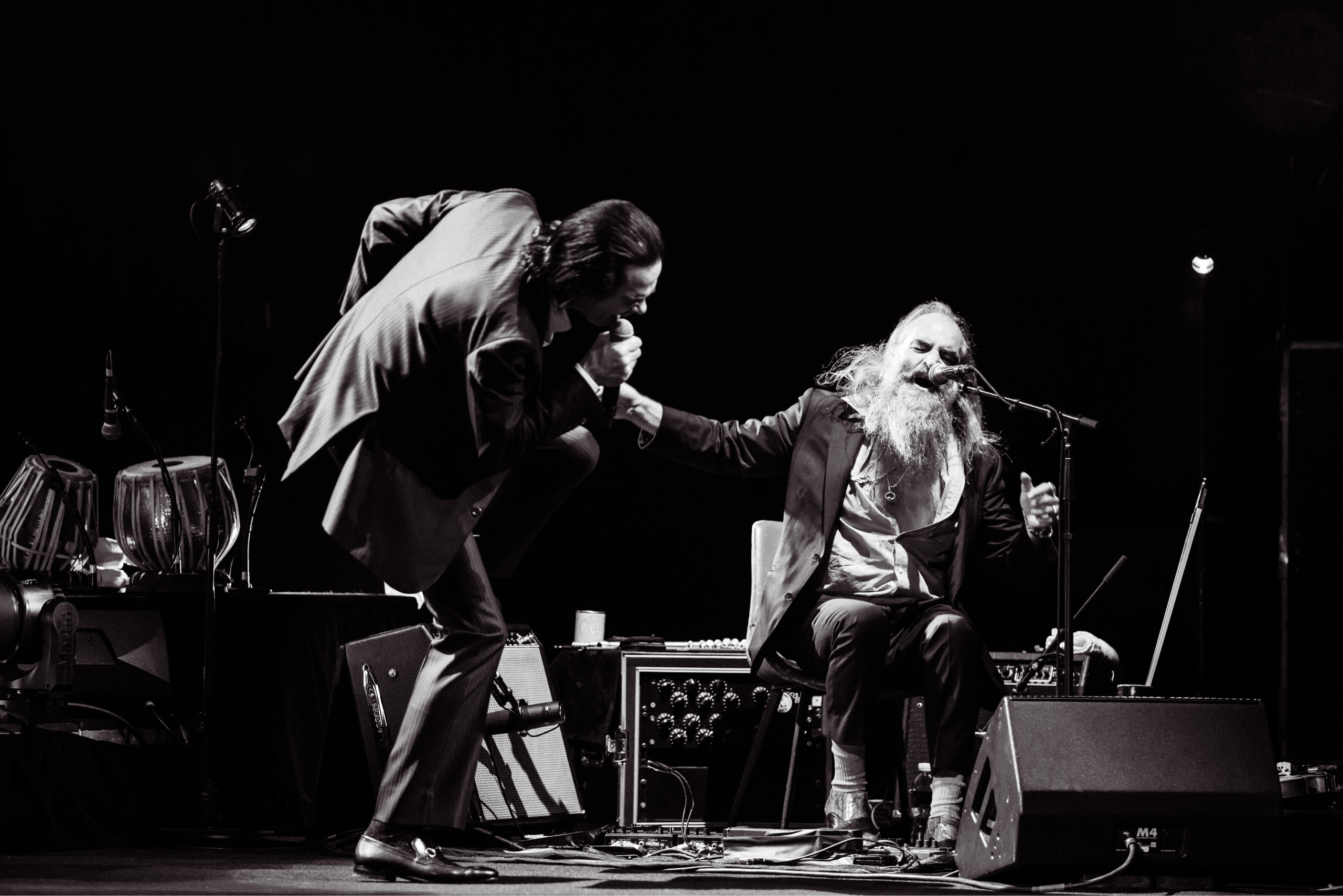 Nick Cave, a tall singer moves energetically during performance while Warren Ellis is seated next to him, singing into a microphone.