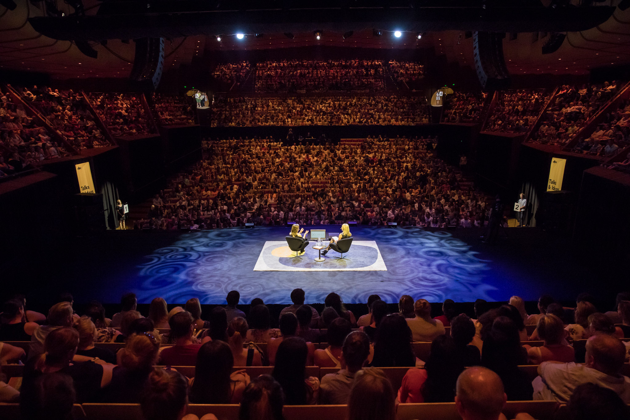 Two people sat on chairs on a stage in the middle of a full seated audience in a red lit theatre.