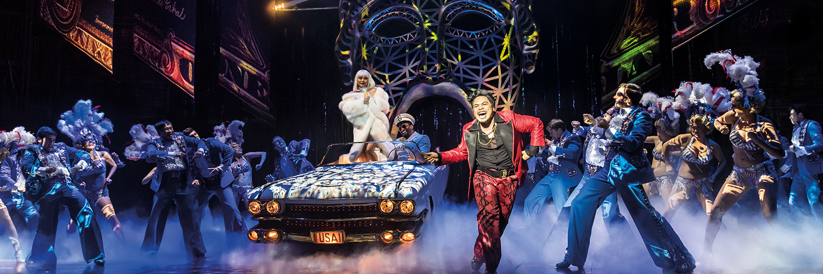 Cast of Miss Saigon on stage, mid-musical number. There are three leads on stage with an ensemble of performers around them.