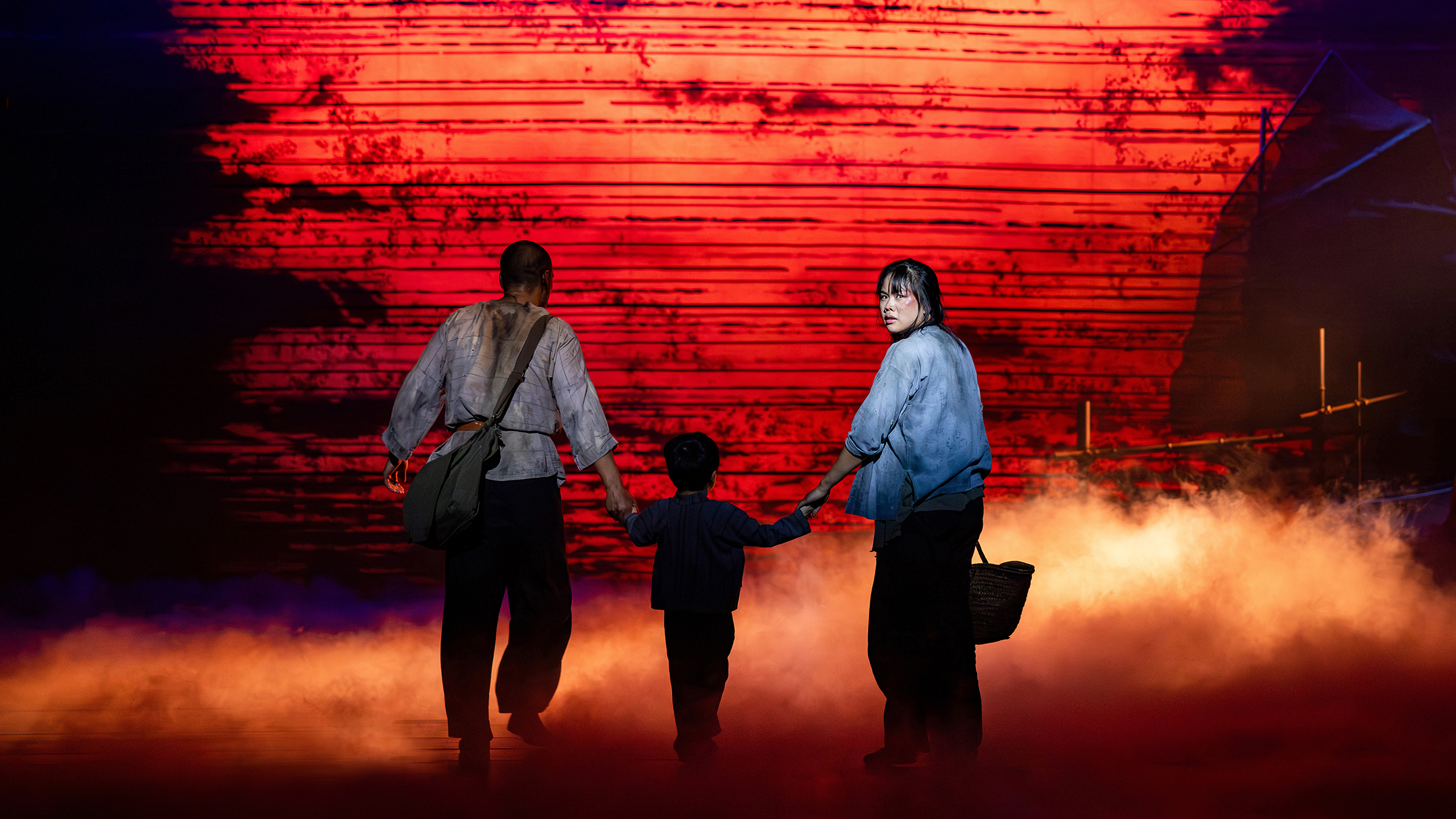 A man wearing a white shirt and dark pants with dirt on it, a small child and an Asian woman wearing a blue shirt with dirt on it and dark pants walk away from the audience. The woman has turned to look at the audience.