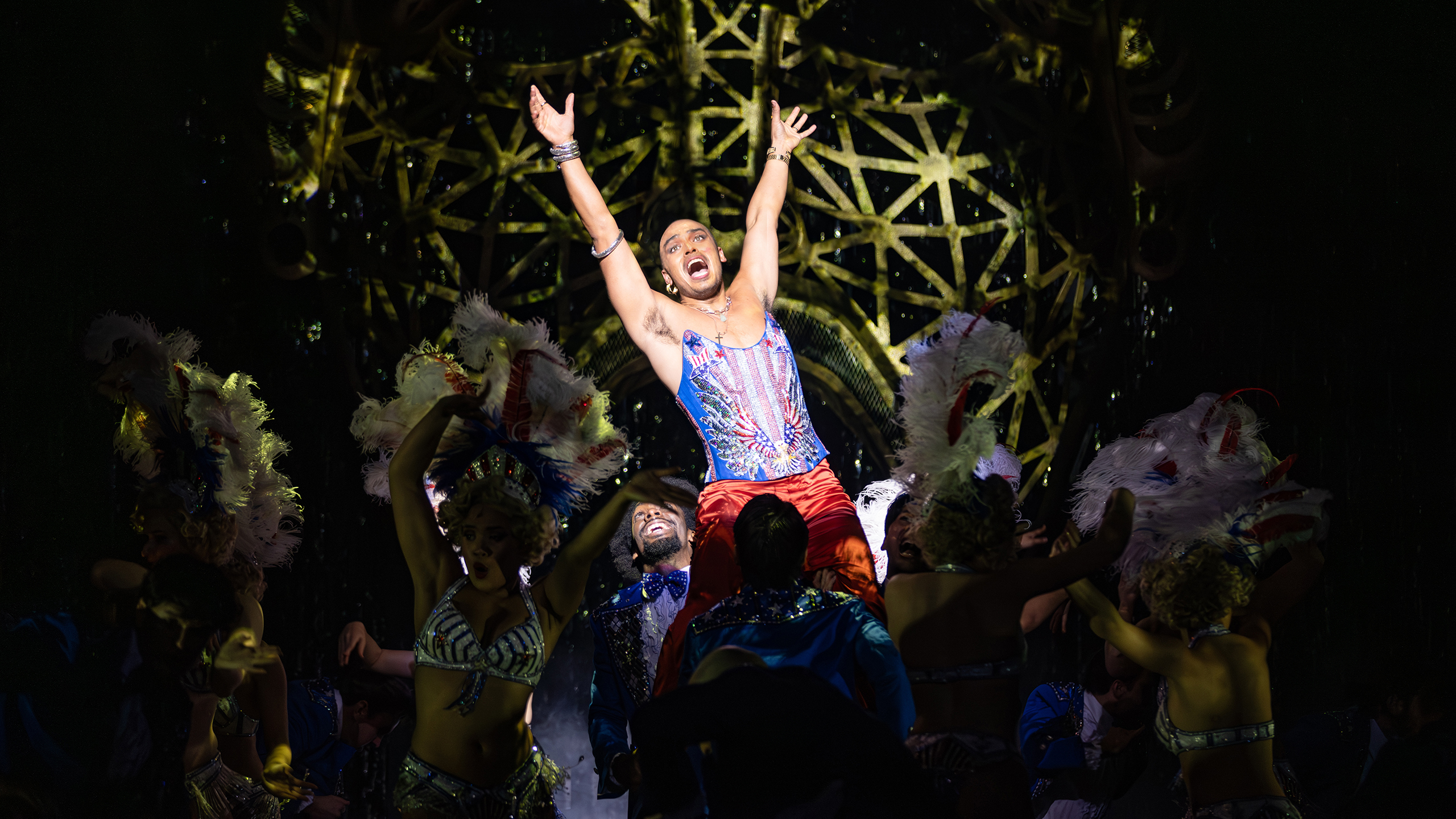 A man in a blue and red sparkly corset stands on top of a car with his arms up.