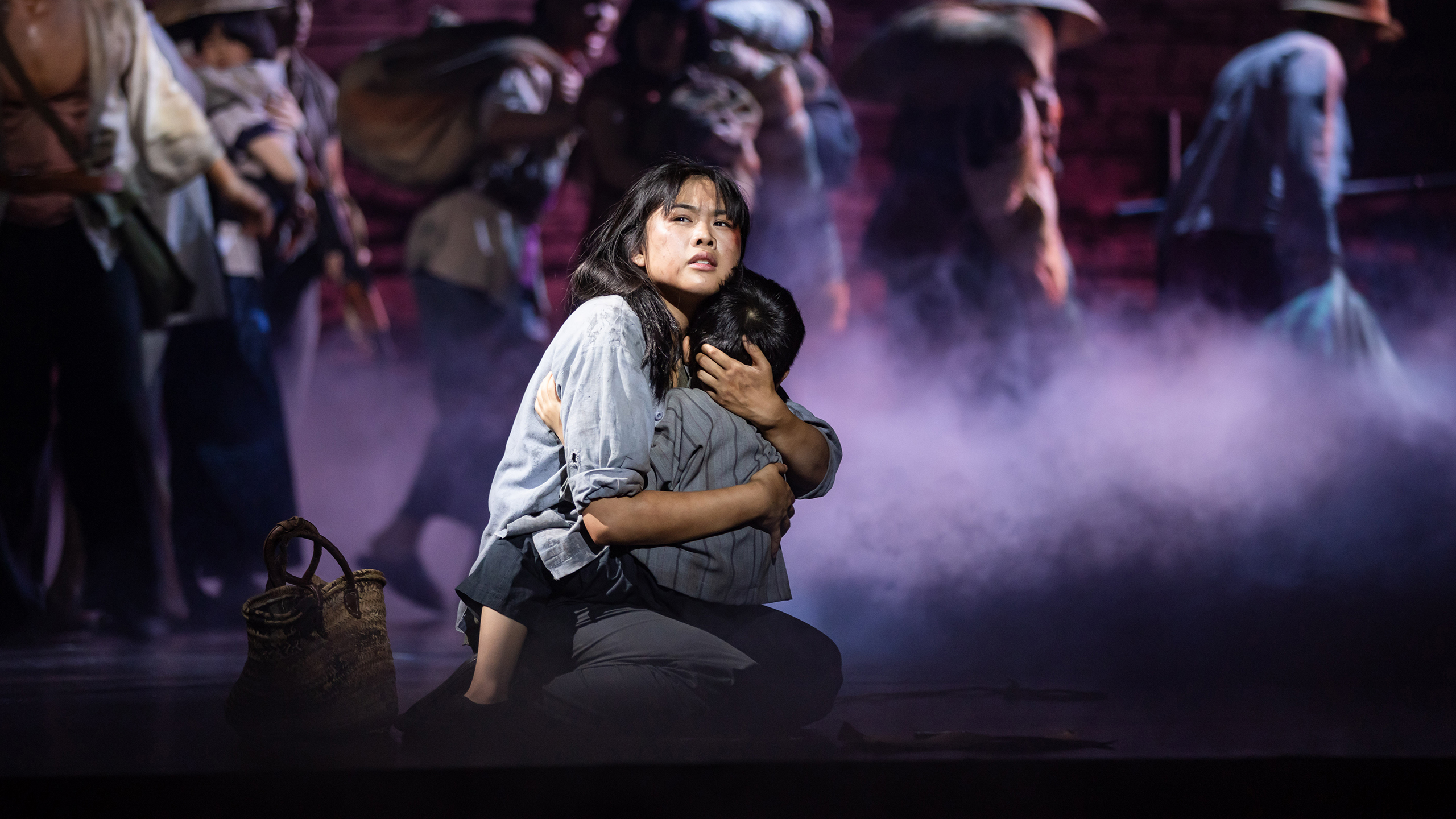 A young Asian woman kneels on stage playing the role of Kim in Miss Saigon, she holds a young child close to her chest and looks solemnly out to the audience.