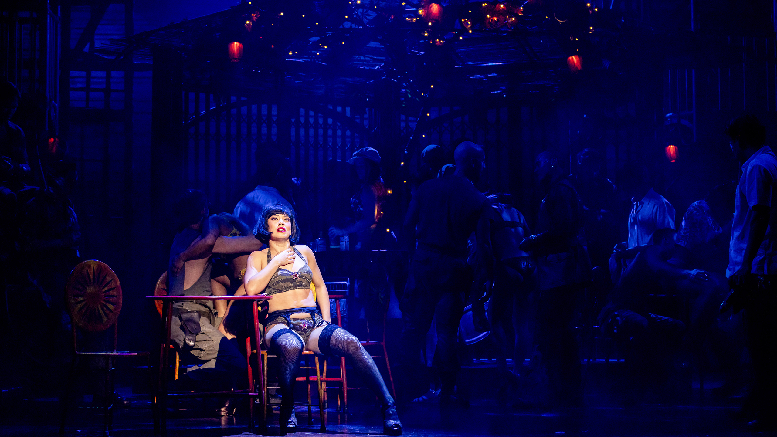 A woman with a short black wig wearing a camouflage coloured bikini sits on a chair with a spotlight on her.