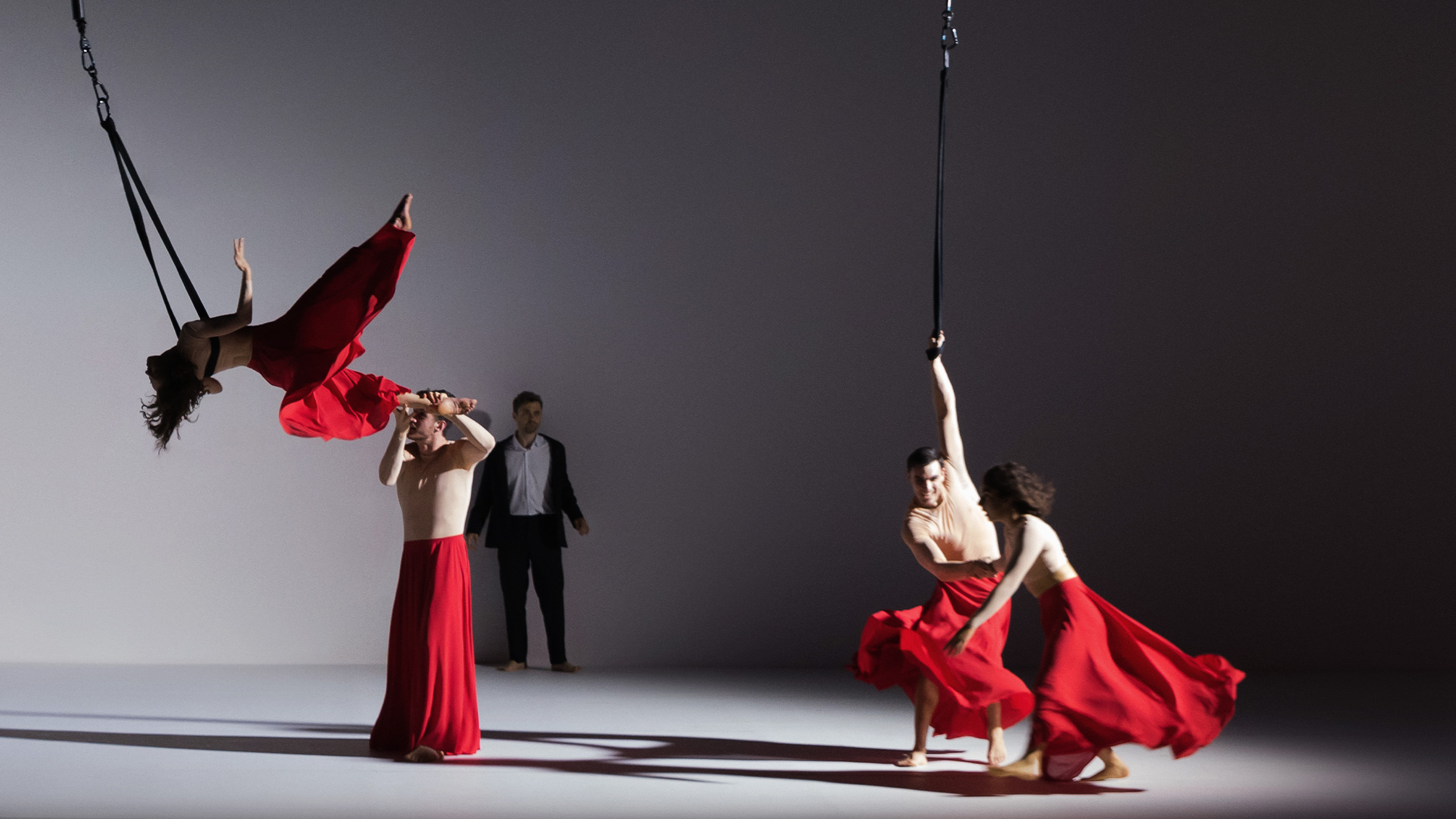 Two cast members wearing long red skirts, one is horizontal in the air attached to an aerial rope, the other assisting her movement
