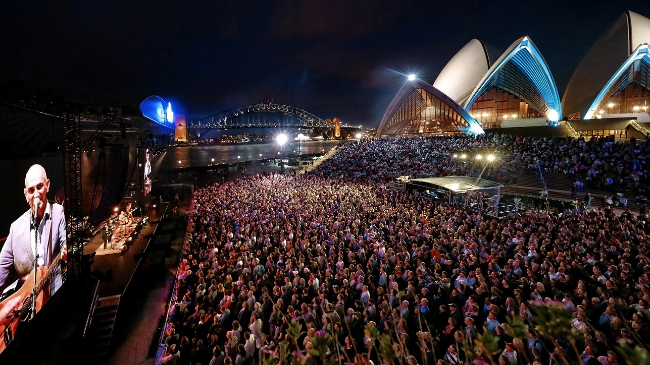 Paul Kelly on stage on the Forecourt playing to a full crowd at night, with the Opera House and the Harbour Bridge in the background.