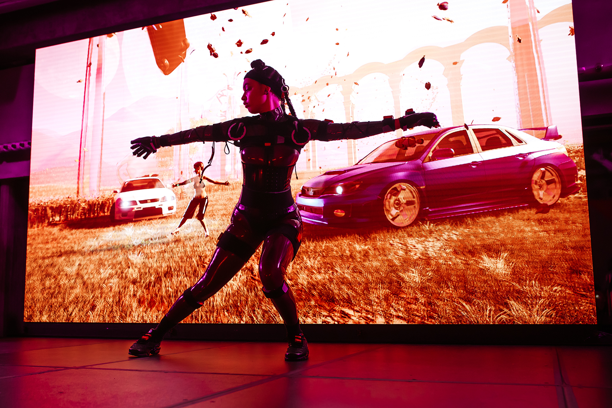 A performer on stage in front of a screen image of two cars in a field and a figure mimicking the movement of the performer