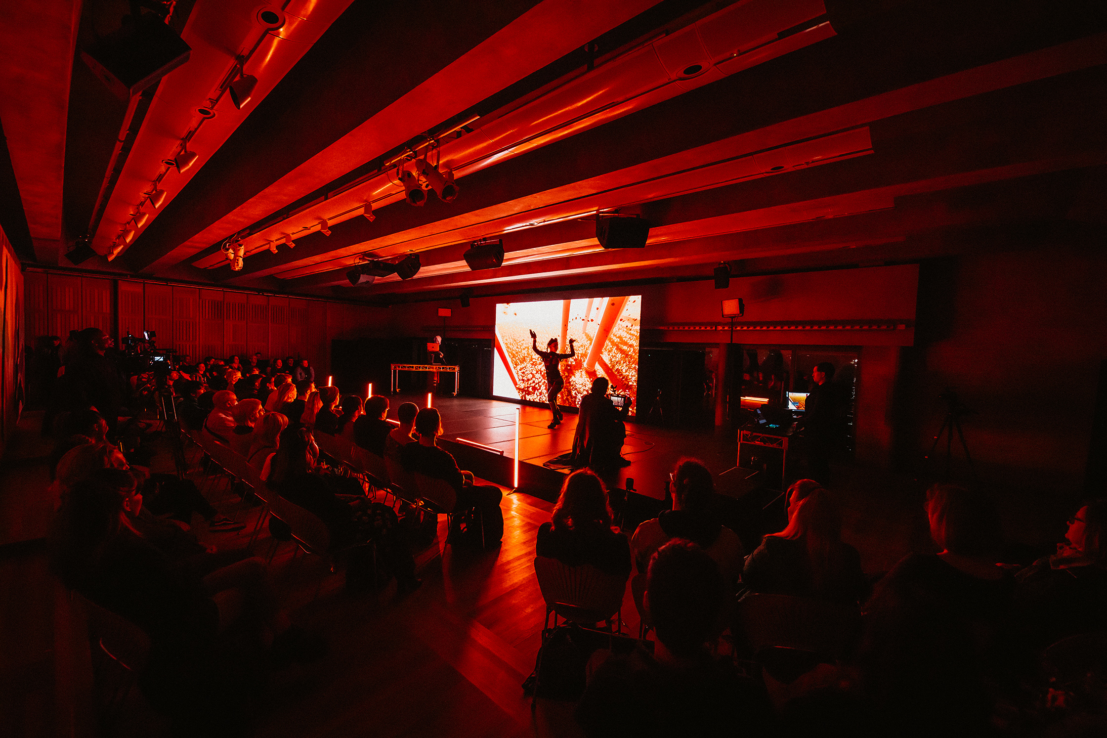 An audience watching a performer in front of a screen image under a dark red light