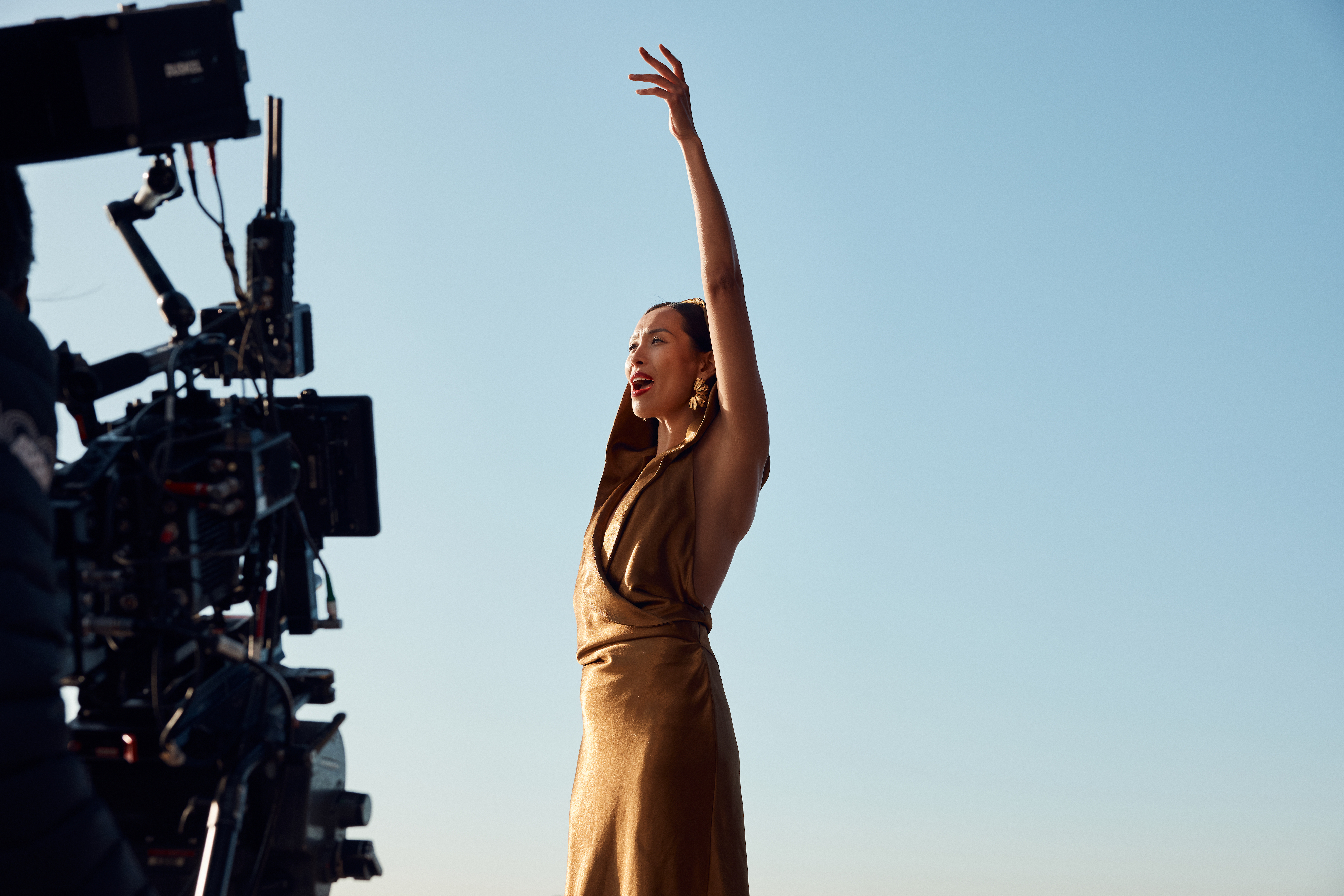 A person wearing a gold dress, signing and holding one hand in the air, a clear blue sky in the background.