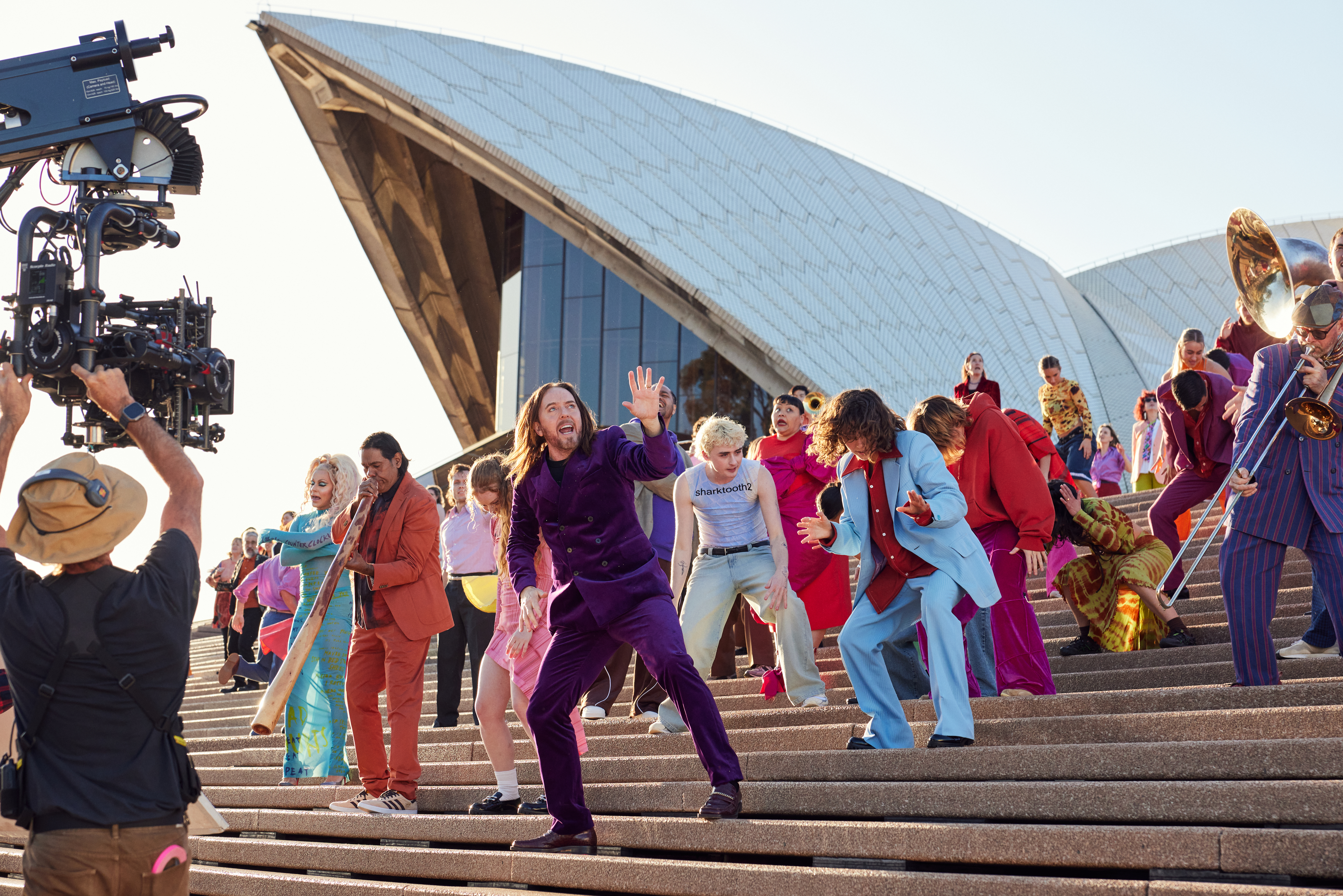 A group of people wearing colourful clothes dancing and playing instruments on the steps of the Sydney Opera House.