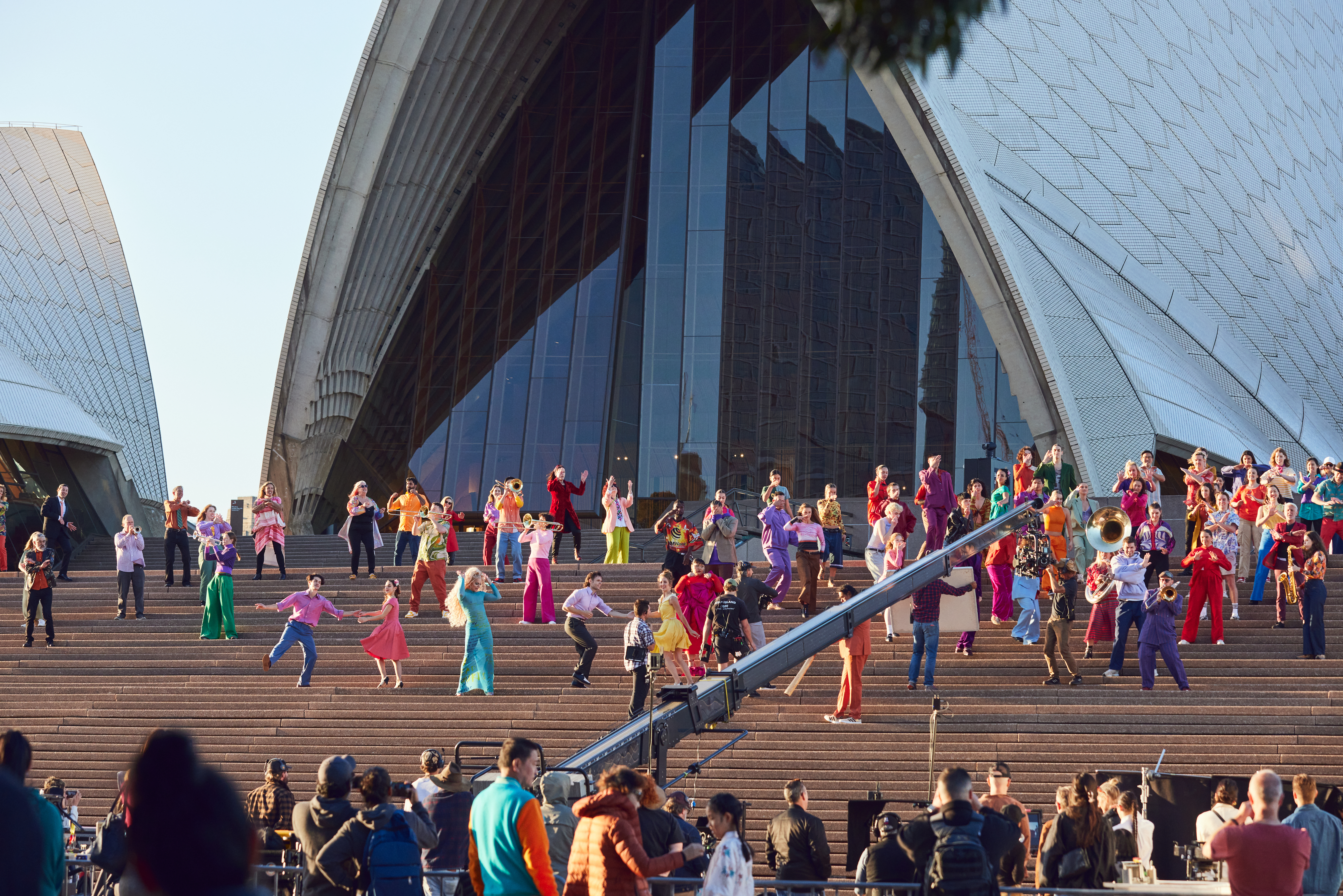A large group of people wearing colourful clothes on the steps of the Sydney Opera House, viewed from a distance.