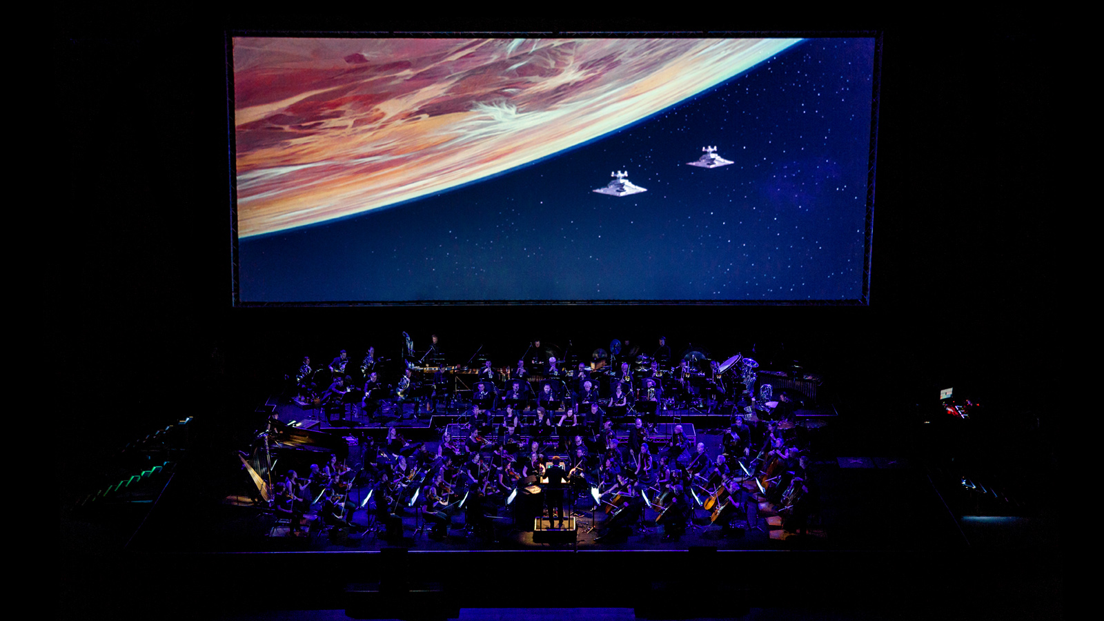 A screen with spaceships and planets on overlooking an orchestra on stage.