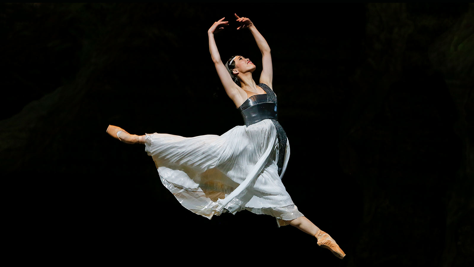 A woman in a white dress leaping with her hands above her head.