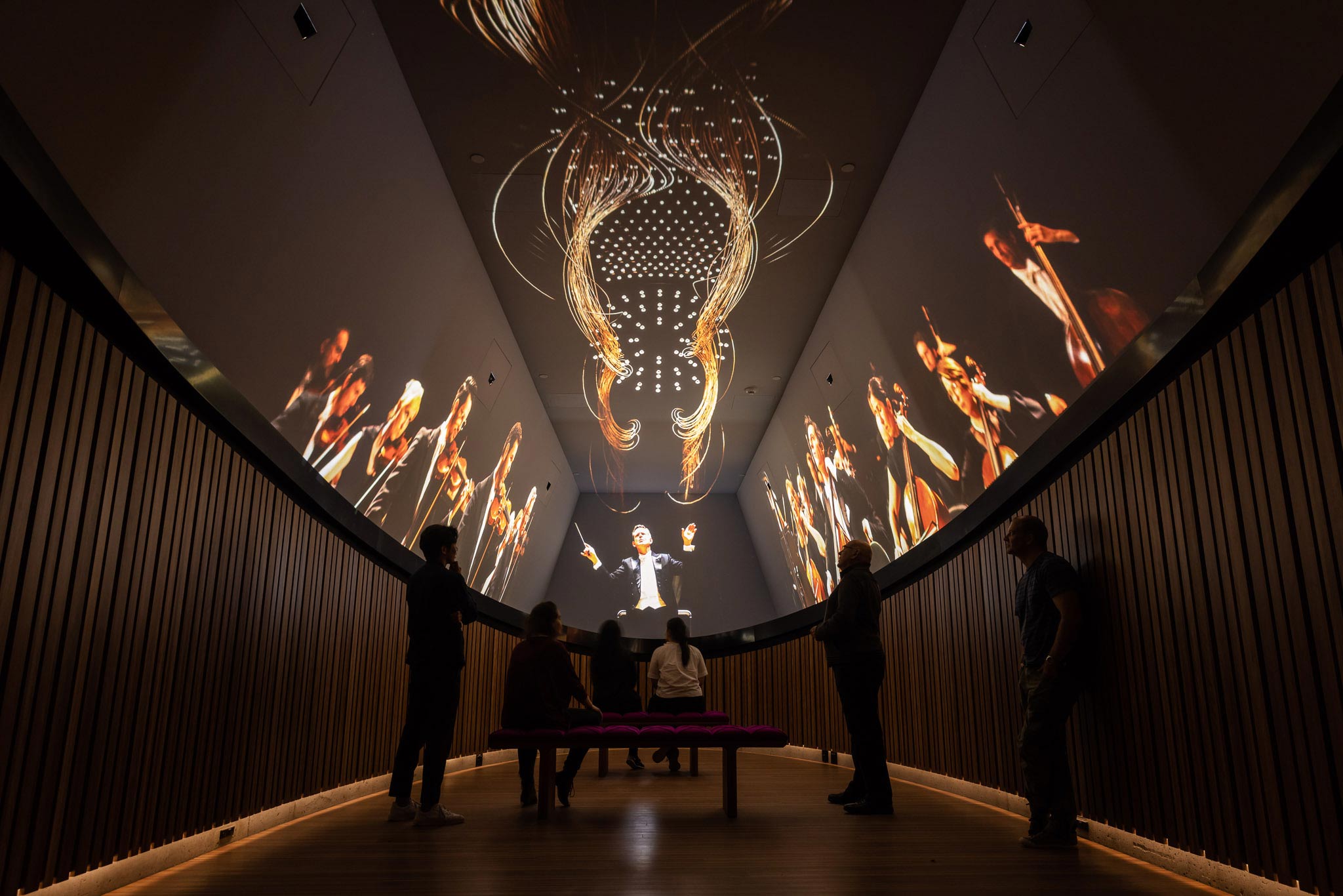 A group of visitors watching a projection of an orchestra show on ceiling screens.