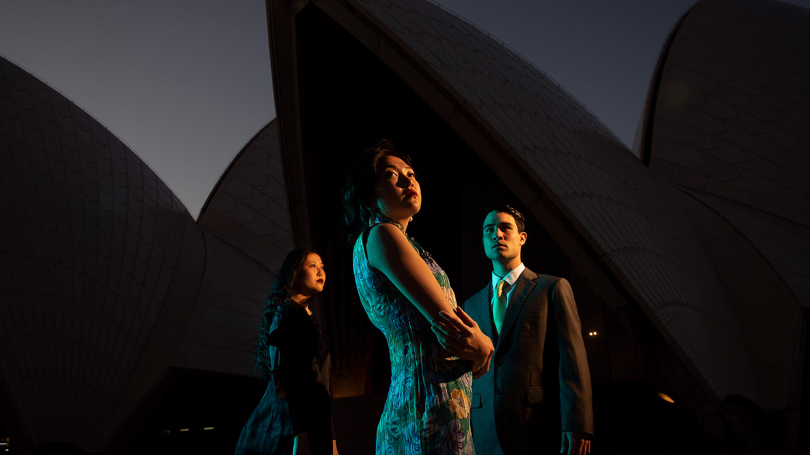 A man in a suit and two women in dresses standing outside the Sydney Opera House.