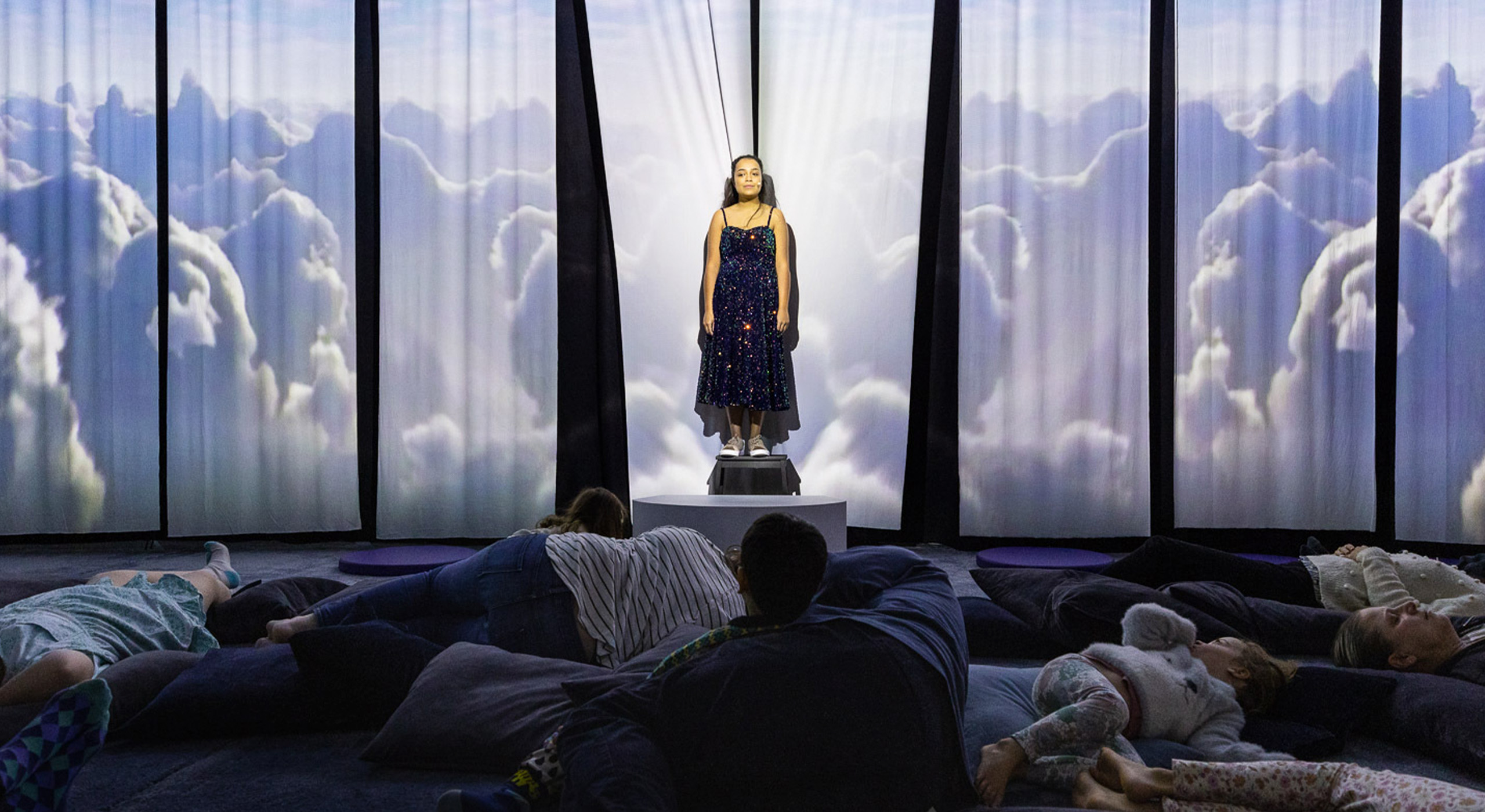 A young white woman with dark brown hair stands in front of a fabric background, where projections of clouds are being cast. An audience of meditation children and adults lie on pillows in front of her.