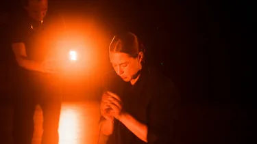 A performer in black kneels on the ground as another performer stands behind her.