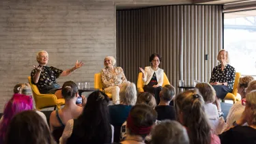 Four women sit on yellow chairs arms aloft.