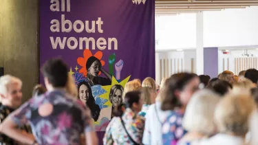 Women stand in a lobby in front of an all about women sign.