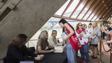 Three women sit at a table signing books in front of a view of the Harbour bridge.