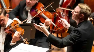 A conductor leads an orchestra as musicians play string instruments, including violins and cellos.