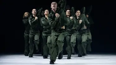 Group of dancers wearing green jumpsuits performing synchronised movements on a stage with a dark background.