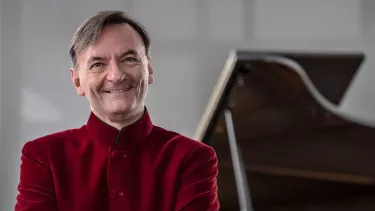 Stephen Hough wears a red shirt standing in front of a grand piano