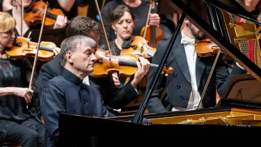 Stephen Hough wears a pinstripe shirt as his eyes are closed playing the piano in front of the violin section of an orchestra