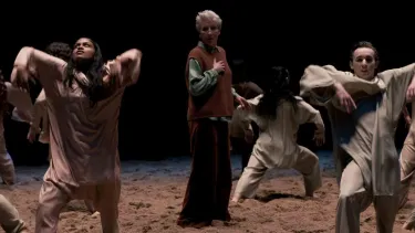 A group of people in loose, earth-toned clothing dance expressively on a sandy stage.