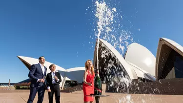 A woman in a red dress and two people in suits standing outside the Sydney opera House, watching a bottle of Champaigne spray into the air.