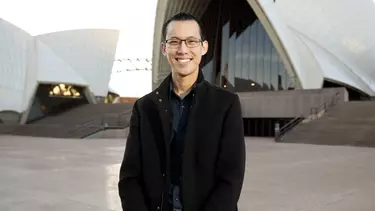 A man in a black jacket and glasses smiling outside of the Sydney Opera House.