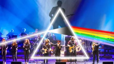 A large band is on a stage lit up in blue. In the foreground a seated crowd is visible. A graphic of a silhouette guitar player, white triangle and rainbow is overlayed.