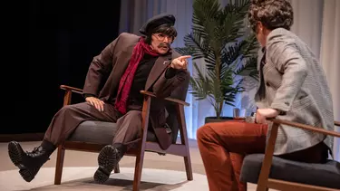 Two actors dressed as men wearing suits and short wigs sit on chairs. They are talking to each other in a scene.