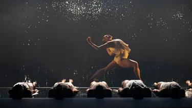 A dancer in a yellow costume performs energetically above five dancers lying on the stage.