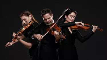 A group of three violinists playing their instruments against a black background.