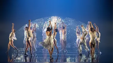 A group of dancers in white costumes performing on stage with arms raised, set against a backdrop of sparkles and dim blue lighting.