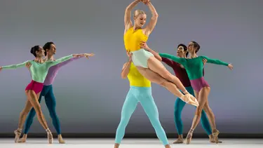 Five ballet dancers on stage: a female dancer in a yellow top being lifted by a male dancer, while three other male dancers in colorful outfits pose around them.