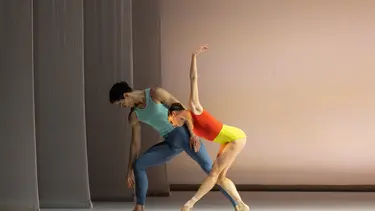 A male ballet dancer stands with his back to a female dancer who leans toward him with one arm raised.