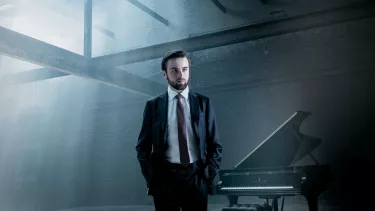 Pianist Daniil Trifonov stands in a suit in front of a grand piano