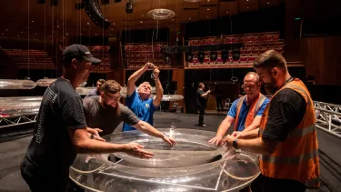 A group of people standing around a perspex disc in the Concert Hall, removing strings from it.