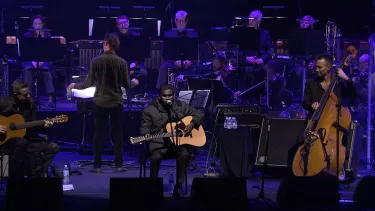 An orchestra on stage under blue lighting, a person playing guitar and singing is highlighted.