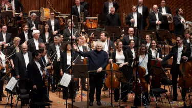 An orchestra on stage, members of the orchestra are standing, the conductor is waving their right arm, smiling.
