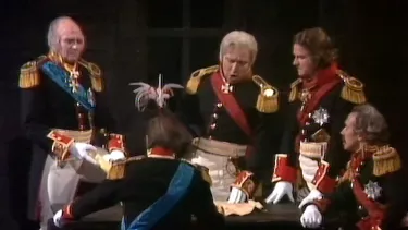 Five people in military dress around a table in conversation. One person is talking with animation, while looking at a paper on the table.