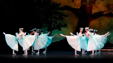 Two groups of six ballerinas on stage, in arabesque, wearing long white tutus and flower crowns.