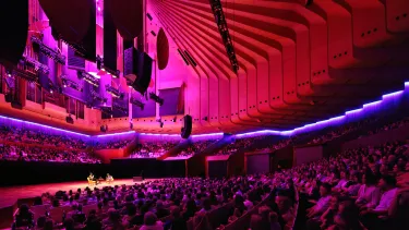 Two speakers on stage with the Concert Hall lit up in red and purple.