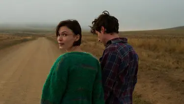 Two individuals stand on a remote dirt road, surrounded by open land and sparse vegetation.