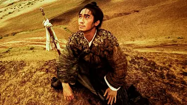 A man is crouched atop a hill in the dessert. He is looking up to the sky and has a sword at his side.