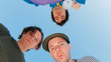 Three people look down at the camera, faces curious and playful. They are outside under a clear blue sky.