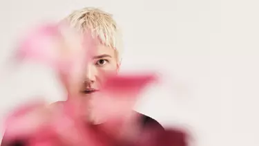 A person with short blonde hair standing behind some pink foliage against a white background.