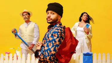 A man stands in the foreground wearing a beanie, blue shirt with flowers on it and a red backpack. Behind him is a white picket fence. Behind the fence to the left is a man wearing a yellow vest and hat and holding a shovel. He looking curiously at the ma
